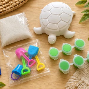 May include: A white ceramic turtle figurine, a clear plastic bag containing white sand, two blue-handled paintbrushes, a clear plastic bag with small colorful plastic shovels and buckets, and two strips of green paint pots for a craft kit.