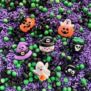 May include: A collection of Halloween-themed resin charms including a black cat in a pumpkin, a ghost in a pumpkin, a purple witch hat, a skull pirate hat, a black cauldron, a white ghost with a broom, and a black spider, set in purple rice.