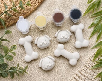 May include: A set of white, unpainted ceramic figures including three dog head shapes and three bone shapes, accompanied by a plastic strip of four small paint pots containing white, yellow, brown, and black paint for crafting projects.