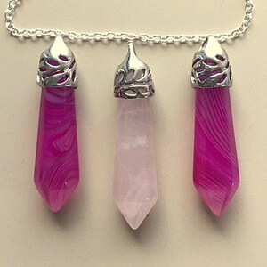 May include: A silver chain necklace with three crystal pendants. The center pendant is a light pink crystal, while the two outer pendants are a darker pink with white swirls. Each pendant is attached to the chain with a silver filigree cap.