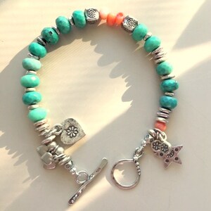 May include: A turquoise beaded bracelet with silver accents and charms. The bracelet features turquoise beads, coral beads, and silver spacers. Charms include a heart with a flower, a star, and other decorative elements. The bracelet has a toggle clasp.