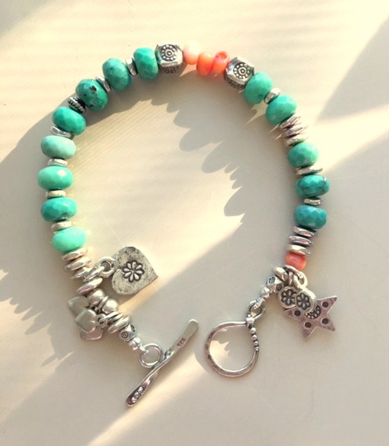 May include: A turquoise beaded bracelet with silver accents and charms. The bracelet features turquoise beads, coral beads, and silver spacers. Charms include a heart with a flower, a star, and other decorative elements. The bracelet has a toggle clasp.