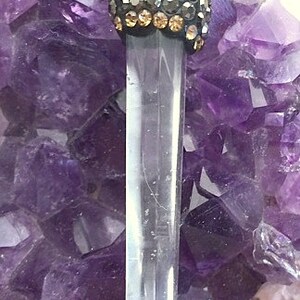 May include: A clear quartz crystal point pendant with a black and gold crystal top. The pendant is displayed on a bed of amethyst crystals.