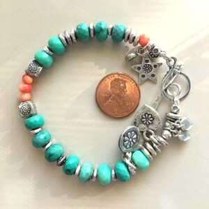 May include: A turquoise beaded bracelet with silver accents and charms, including a star, heart, and flower. The bracelet also features coral-colored beads and is shown next to a penny for size reference.