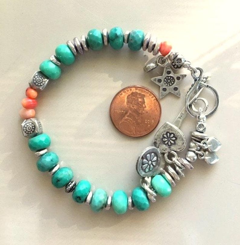 May include: A turquoise beaded bracelet with silver accents and charms, including a star, heart, and flower. The bracelet also features coral-colored beads and is shown next to a penny for size reference.