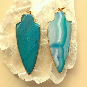 May include: Two blue agate arrowhead pendants with gold-toned metal settings. The arrowheads have a white and blue agate pattern.