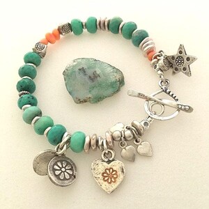 May include: A turquoise beaded bracelet with silver accents, coral beads, and a toggle clasp. The bracelet features a star charm, heart charms, and a flower charm. A rough-cut green stone is next to the bracelet.