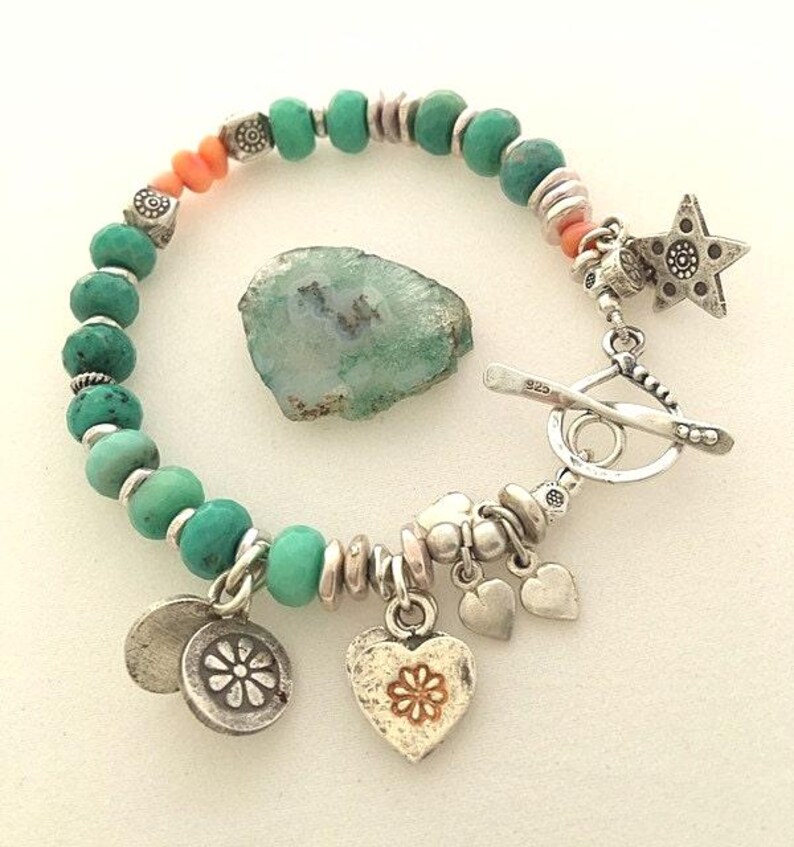 May include: A turquoise beaded bracelet with silver accents, coral beads, and a toggle clasp. The bracelet features a star charm, heart charms, and a flower charm. A rough-cut green stone is next to the bracelet.