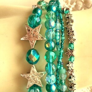 May include: A set of five turquoise and silver beaded bracelets with star charms. The bracelets are layered on a white surface.