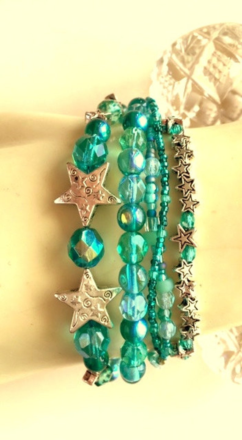 May include: A set of five turquoise and silver beaded bracelets with star charms. The bracelets are layered on a white surface.