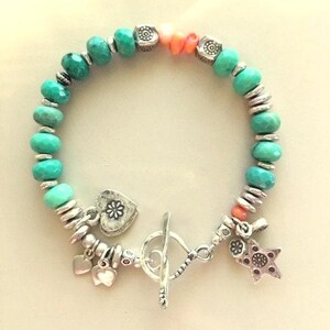 May include: A turquoise beaded bracelet with silver accents. The bracelet features a toggle clasp, heart and star charms, and coral-colored beads. The beads are a mix of round and faceted shapes, creating a textured look.