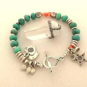 May include: A turquoise beaded bracelet with silver accents, charms, and a toggle clasp. The bracelet features a clear quartz crystal pendant. Charms include hearts and a star.