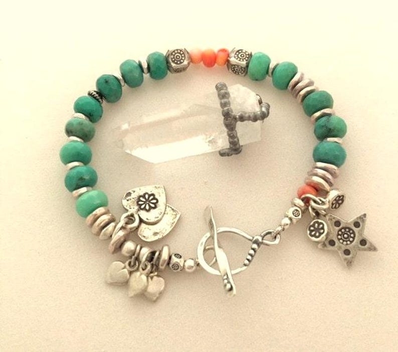 May include: A turquoise beaded bracelet with silver accents, charms, and a toggle clasp. The bracelet features a clear quartz crystal pendant. Charms include hearts and a star.