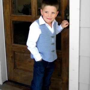 Op de afbeelding: Een jonge jongen draagt een lichtblauwe gehaakte vest over een wit overhemd met knopen en een blauwe jeans. Hij staat voor een houten deur met een messing deurknop.
