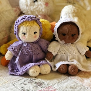Könnte beinhalten: Zwei gestrickte Puppen, eine mit einem lila Kleid und einer gelben Haube, die andere mit einem weißen Kleid und einer braunen Haube. Die Puppen sitzen auf einer Patchwork-Decke.