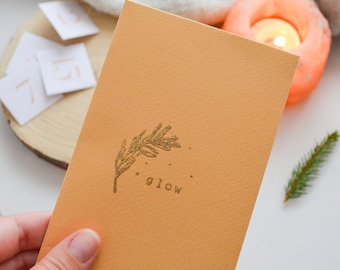 May include: A mustard-colored card with a gold glitter sprig and the word "glow" printed on it. The card is held in a hand, with a lit candle and small numbered cards in the background. A sprig of greenery is also present.