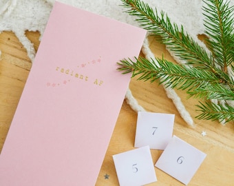 May include: A pink card with the text "radiant AF" in gold lettering, accompanied by three small white cards numbered 5, 6, and 7. The items are arranged on a wooden surface with a sprig of evergreen and a white, textured fabric.