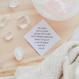 May include: A white card with the text "Do some star or cloud gazing. If it's cold add a blanket and a hot drink. Let go of your thoughts and just watch." on a wooden surface with white stones and a white blanket.