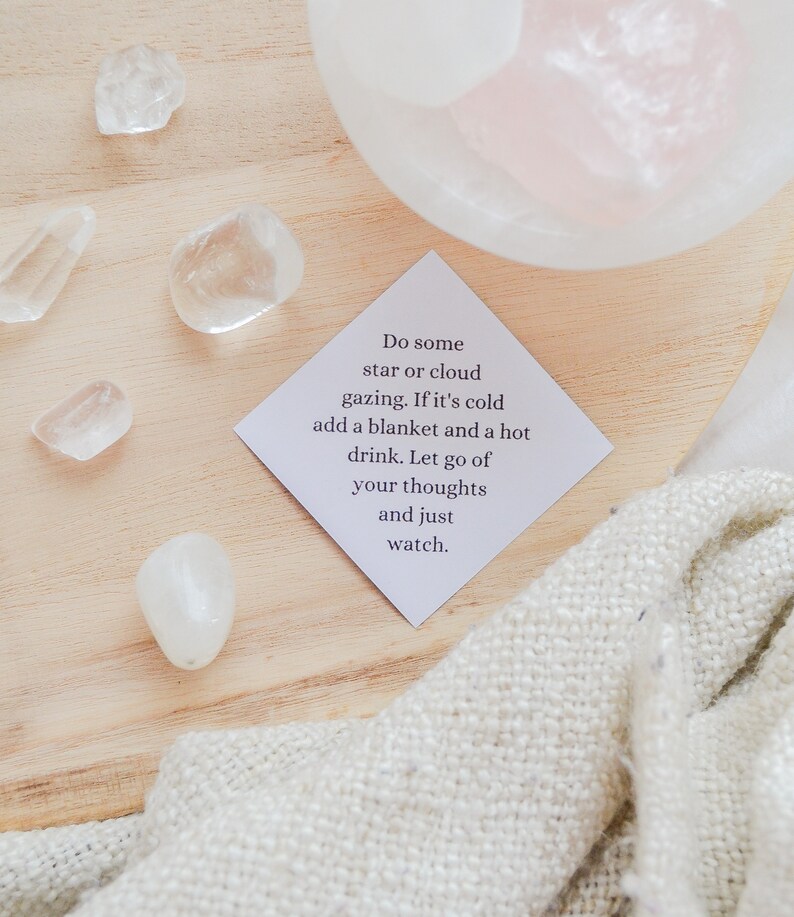 May include: A white card with the text "Do some star or cloud gazing. If it's cold add a blanket and a hot drink. Let go of your thoughts and just watch." on a wooden surface with white stones and a white blanket.