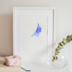 May include: A framed watercolor painting of a blue crescent moon. The artwork is displayed in a white frame and rests on a white surface. A pink crystal and a glass vase with greenery are also on the surface.