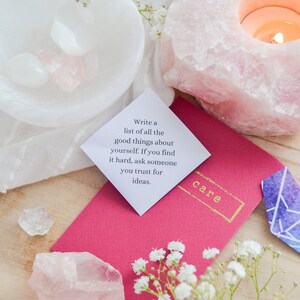 May include: A pink and white crystal with a candle inside, a white bowl with crystals, a pink envelope with the words "Write a list of all the good things about yourself. If you find it hard, ask someone you trust for ideas." and the word "care" in gold lettering, and a small blue and purple crystal.