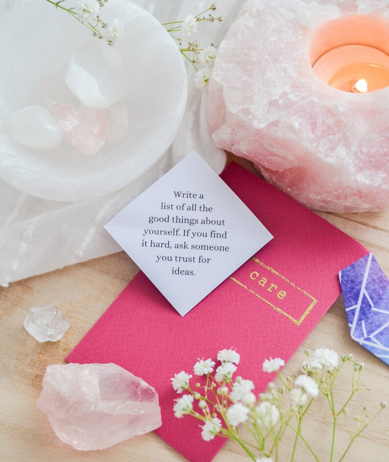 May include: A pink and white crystal with a candle inside, a white bowl with crystals, a pink envelope with the words "Write a list of all the good things about yourself. If you find it hard, ask someone you trust for ideas." and the word "care" in gold lettering, and a small blue and purple crystal.