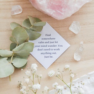 May include: A white card with the text "Find somewhere calm and just let your mind wander. You don't need to work anything out. Just be." on a wooden surface with white flowers, green leaves, and clear quartz crystals.