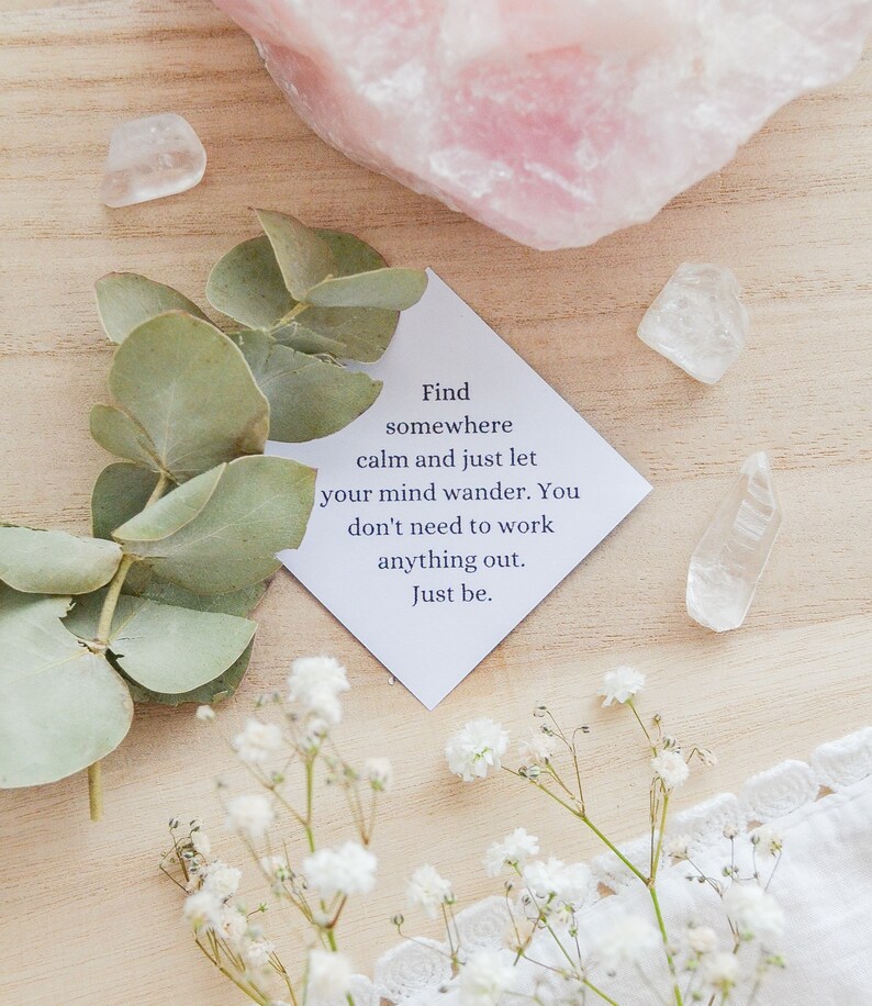 May include: A white card with the text "Find somewhere calm and just let your mind wander. You don't need to work anything out. Just be." on a wooden surface with white flowers, green leaves, and clear quartz crystals.