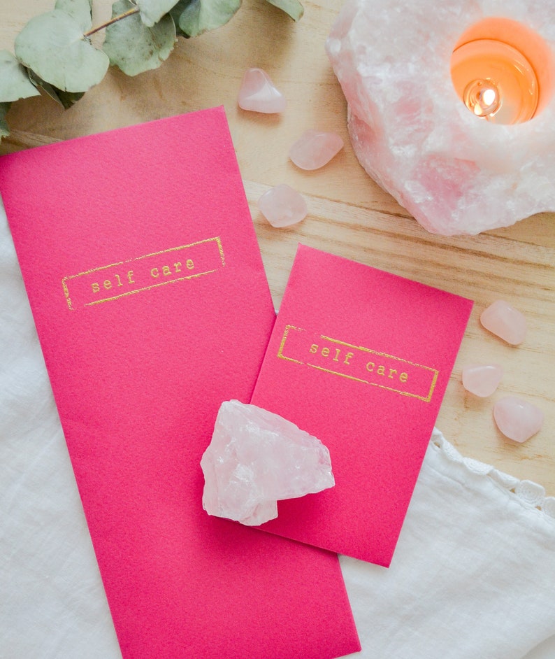 May include: Two pink cards with the text "self care" in gold lettering on a wooden surface with pink crystals and a lit candle.