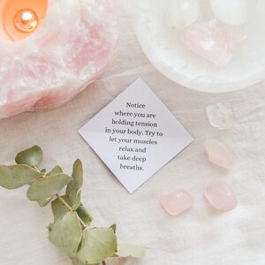 May include: A white card with the text "Notice where you are holding tension in your body. Try to let your muscles relax and take deep breaths."  The card is on a white fabric surface with pink rose quartz crystals and a sprig of eucalyptus.
