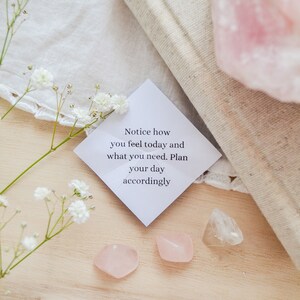 May include: A white card with the text "Notice how you feel today and what you need. Plan your day accordingly."  The card is surrounded by white flowers, pink and clear crystals, and a white cloth.