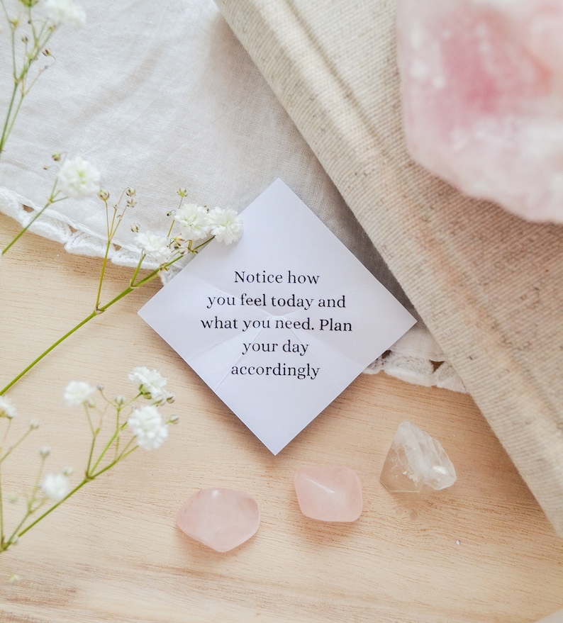 May include: A white card with the text "Notice how you feel today and what you need. Plan your day accordingly."  The card is surrounded by white flowers, pink and clear crystals, and a white cloth.