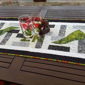 May include: A black and white quilted table runner with a green leaf design. The runner has a red and green border and is decorated with two clear glasses and a wooden sculpture.
