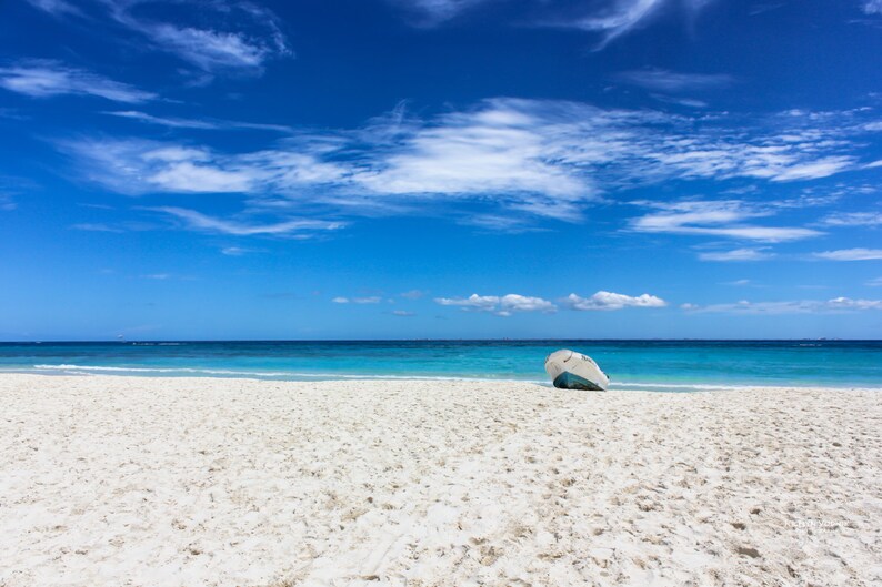 May include: A bright, sunny beach scene with a clear blue sky dotted with fluffy white clouds. The turquoise ocean meets a white sandy beach. A small, weathered boat rests on the sand, adding a touch of nautical charm to the serene landscape.