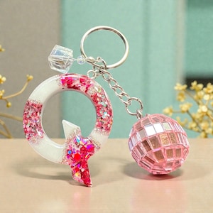 May include: A keychain featuring a white and glittery pink letter "Q" pendant and a small pink disco ball charm. The letter is accented with red glitter and a clear crystal bead. The keychain is attached to a silver-colored ring.