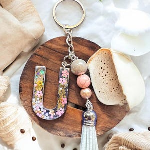 May include: A white tassel keychain with a silver charm and pink and beige beads. The charm is a clear resin letter U with glitter and pink flowers.