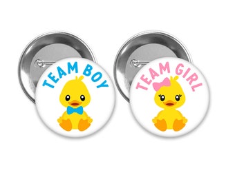 May include: Two round buttons with a white background and silver metal backing. The button on the left has a yellow duckling with a blue bow tie and the text "Team Boy". The button on the right has a yellow duckling with a pink bow and the text "Team Girl".