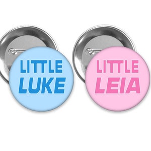 May include: Two round button pins with silver backing. The left pin is light blue with the text "LITTLE LUKE" in blue. The right pin is pink with the text "LITTLE LEIA" in pink.