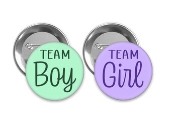 May include: Two round buttons with silver backs. The button on the left is light green with the text "TEAM Boy" in black. The button on the right is light purple with the text "TEAM Girl" in purple.