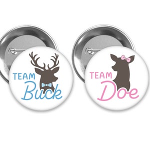 May include: Two round buttons with a white background and silver trim. The left button features a brown deer silhouette with antlers and a blue bow tie. The text "TEAM BUCK" is written in blue cursive. The right button features a brown deer silhouette with a pink bow. The text "TEAM DOE" is written in pink cursive.