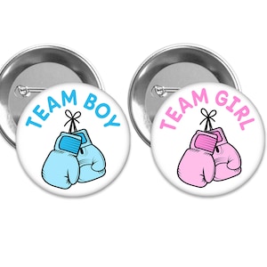 May include: Two round white buttons with silver backing. The button on the left says "Team Boy" in blue text with a blue boxing glove illustration. The button on the right says "Team Girl" in pink text with a pink boxing glove illustration.
