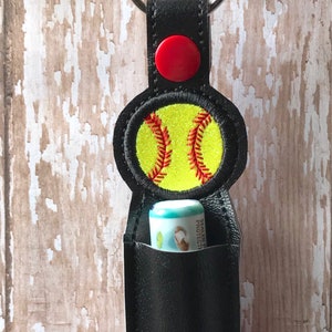 May include: Black leather keychain with a red button and a yellow softball design. The keychain has a pocket for a lip balm tube.