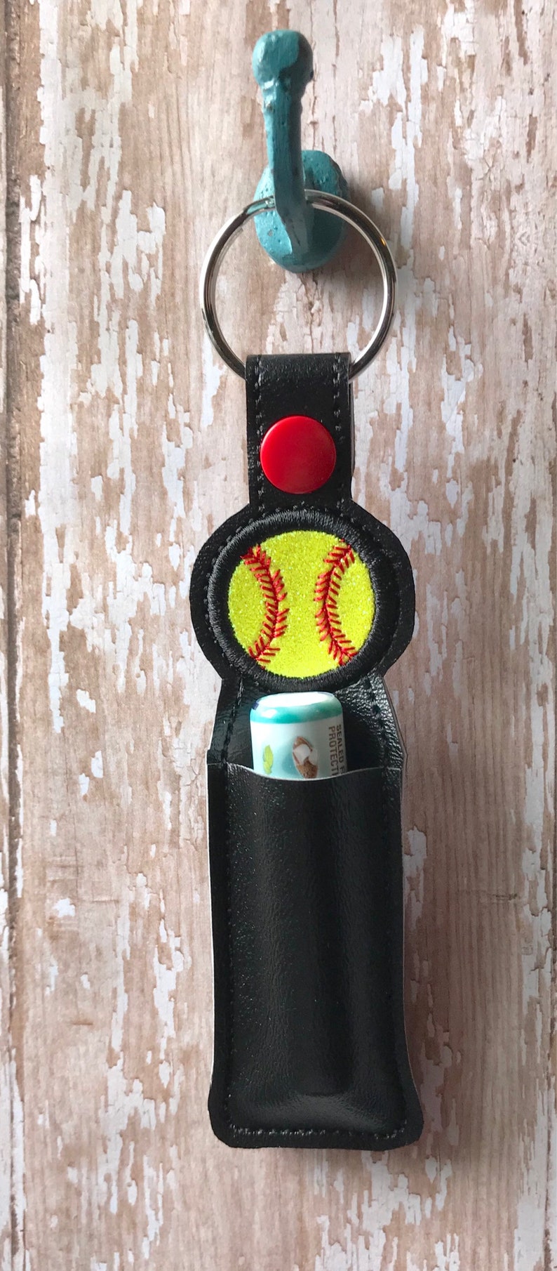 May include: Black leather keychain with a red button and a yellow softball design. The keychain has a pocket for a lip balm tube.
