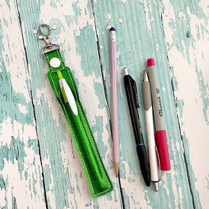 May include: A green fabric pen holder with a silver keychain. The holder is lying on a light blue wooden surface with a pink pencil, a black pen, and a silver pen with a red marker.