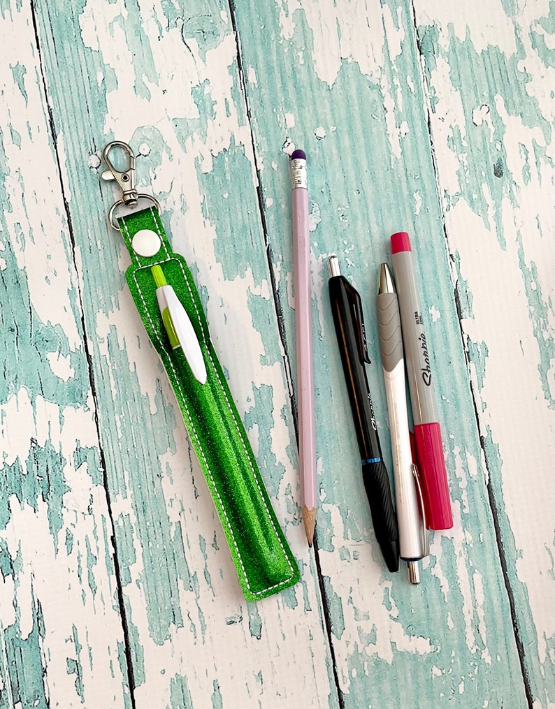 May include: A green fabric pen holder with a silver keychain. The holder is lying on a light blue wooden surface with a pink pencil, a black pen, and a silver pen with a red marker.