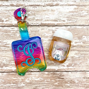 May include: A rainbow-colored hand sanitizer holder with a blue letter "G" monogram. The holder has a silver key ring and is attached to a small bottle of Pocketbac hand sanitizer with the scent "Toasted Vanilla Chai".