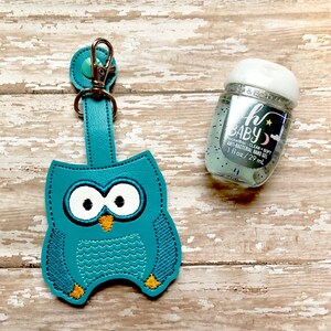 May include: Teal blue owl-shaped hand sanitizer holder with a silver clip and a small bottle of clear hand sanitizer with a white cap and the text "Baby PocketBac Clean + Soft Anti-Bacterial Hand Gel 1 fl oz/29 mL"