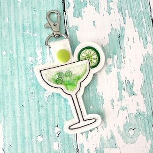 May include: White fabric keychain with a green margarita glass and lime wedge. The glass is filled with a green liquid and has a black outline.