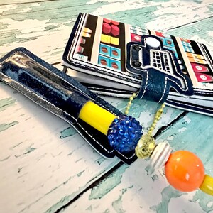May include: A blue denim pen holder with a yellow pen and a blue rhinestone charm. The pen holder is attached to a small notebook with a colorful design of a grocery store.