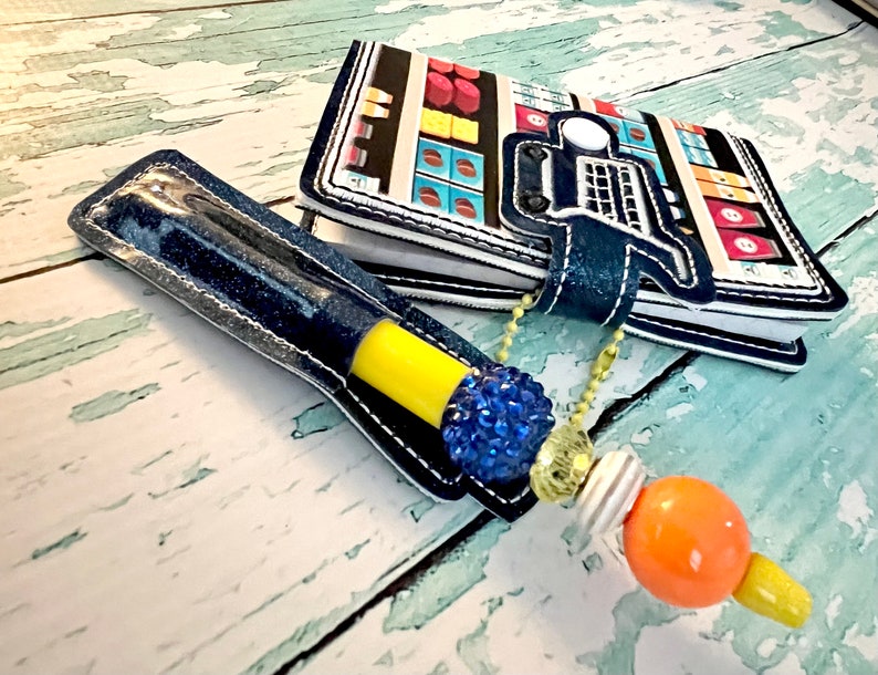 May include: A blue denim pen holder with a yellow pen and a blue rhinestone charm. The pen holder is attached to a small notebook with a colorful design of a grocery store.